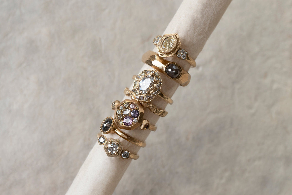 A stack of seven vintage-style gold rings with various gemstone settings, displayed on a cream-colored ring stand against a textured light gray background.